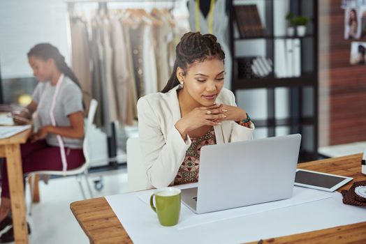 Cropped shot of a young fashion designer working on her laptop with a colleague in the background.