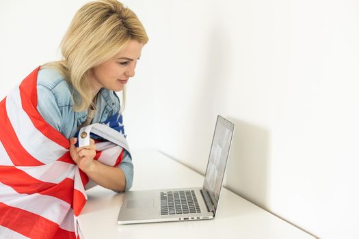 Happy woman employee sitting wrapped in USA flag, shouting for joy in office workplace, celebrating labor day or US Independence day. Indoor studio studio shot isolated on yellow background.