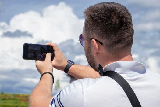 guy with his back turned makes a selfie on the phone. A bearded man in a white polo shirt with a smartphone in his hand takes a picture of himself against the backdrop of a cloudy sky. back view