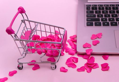 mini shopping basket with red hearts on a pink background. Hearts are scattered over the keyboard of a pink laptop. Love concept of ordering gifts, shopping online. High quality photo