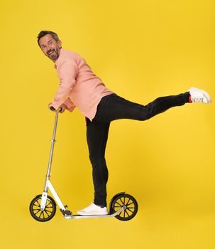 Middle aged grey haired man ride scooter stretched one leg up looking at camera wearing shirt and jeans isolated on yellow background. Handsome mature businessman on the go.