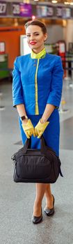 Full length of woman stewardess in aviation air hostess uniform looking at camera and smiling while holding bag for traveling