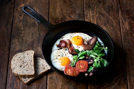 Breakfast set. Pan of fried eggs with bacon, fresh tomato, sage and bread on dark serving board over black wooden background, top view, copy space