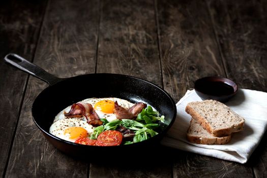 Breakfast set. Pan of fried eggs with bacon, fresh tomato, sage and bread on dark serving board over black wooden background, top view, copy space