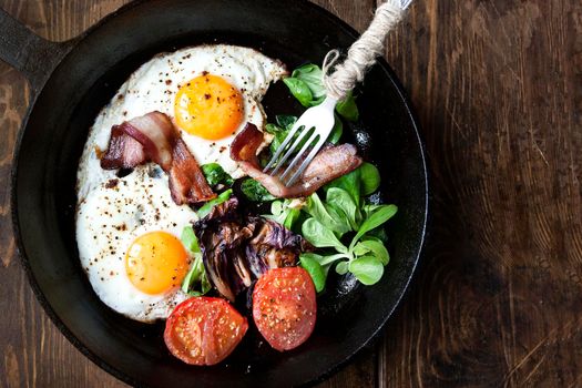 Breakfast set. Pan of fried eggs with bacon, fresh tomato, sage and bread on dark serving board over black wooden background, top view, copy space