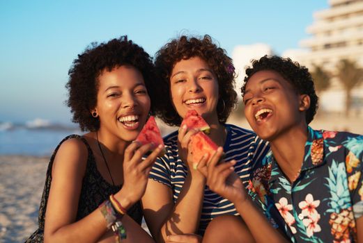 Portrait of three attractive young women eating watermelon pieces on the beach during the day.