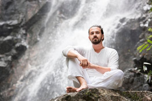 Wellness spa, vacation and yoga meditation concept. Young man sitting in lotus position on the rock under tropical waterfall.