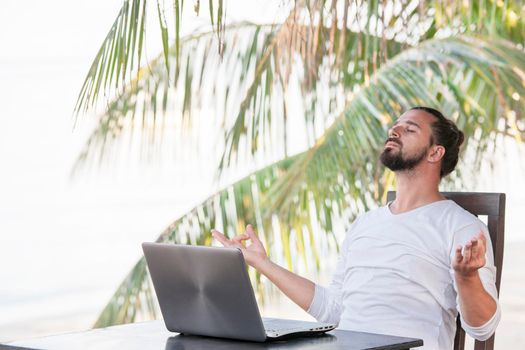 Vacation and technology. Work and travel. Young bearded man using laptop computer while sitting at beach cafe bar near palms and doing yoga