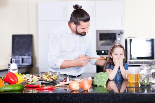 Picture of little girl refusing to eat a salad while cooking and having lunch together with father in the kitchen