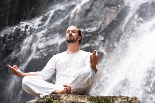 Wellness spa, vacation and yoga meditation concept. Young man sitting in lotus position on the rock under tropical waterfall.