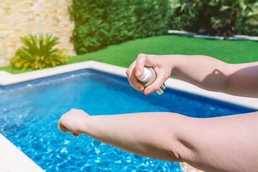 close-up of a young woman's arm applying sunscreen to her skin next to a swimming pool. cosmetic to prevent sunburn. concept of health and skin care. sunlight, outside a garden.