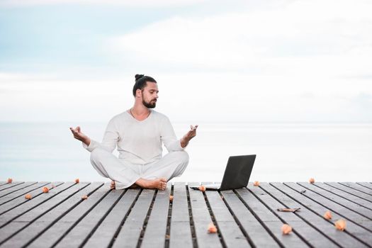 Man in white clothes meditating yoga on wooden pier near ocean