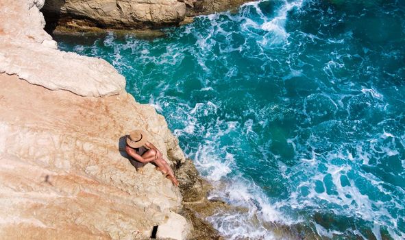 Beautiful young woman sitting on high rock and looking to sea. top view. Girl in Hat on the edge of cliff. blue sea and high cliffs. Seascape. Girl at sunset. Sea tour. Mediterranean Sea, Cyprus.