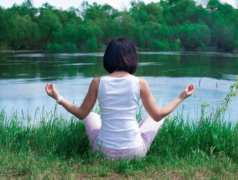 a girl sits in the grass by the river with her back in a meditate pose before. High quality photo. Yoga Concept. Hand Woman Practicing Lotus Pose. High quality photo