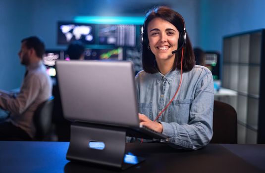 Smiling young female caucasian financial analyst working in the office with laptop and headset looking at camera. Stock market and tradning concept.
