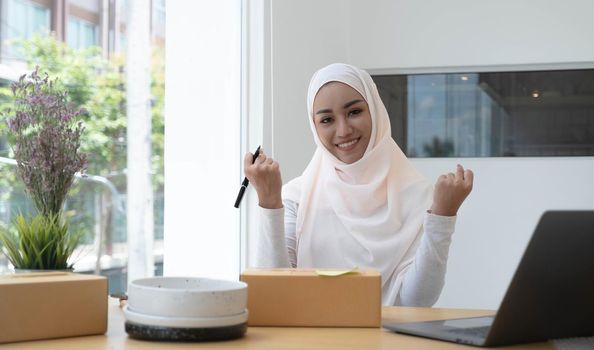 Portrait of a cute smiling Muslim woman wearing a light pink hijab sitting on an office chair. Looking at the camera is delighted while being able to sell online and the background of the box..