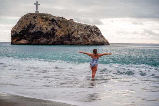 A plump woman in a bathing suit enters the water during the surf. Alone on the beach, Gray sky in the clouds, swimming in winter