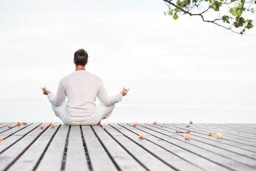 Man in white clothes meditating yoga on wooden pier near ocean