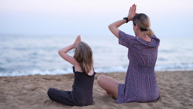 Mom and daughter doing yoga and sitting in lotus position on seashore back view. Pilates sports training with kids concept