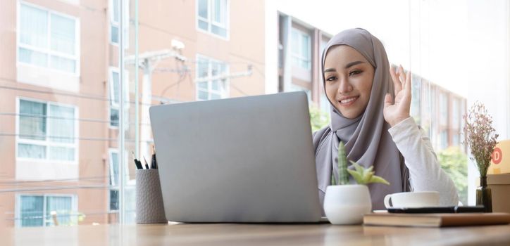 smiling beautiful muslim businesswoman using a laptop for online video meeting sitting at the desk in modern office space, looks at the webcam and waving hello.