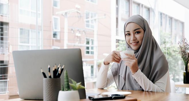 Attractive and cheerful asian muslim female worker or female college student with hijab remote working at the coffee shop, holding a coffee cup..