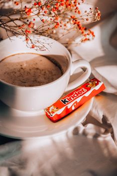 Kiev, Ukraine - 21 January 2021 White cup of milk and KINDER chocolate. Morning aesthetics vibes. Breakfast. Pink gypsophila flowers. Hard shadows.