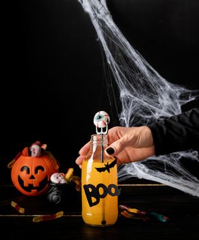 Happy Halloween concept. Woman hand with black nails holding scary colorful Halloween cocktail with party decorations on dark background
