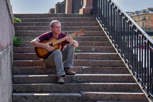 An adult elderly man of retirement age plays an old six-string classical acoustic guitar outdoors while sitting on a granite staircase on a summer evening in the city. Selective focus.