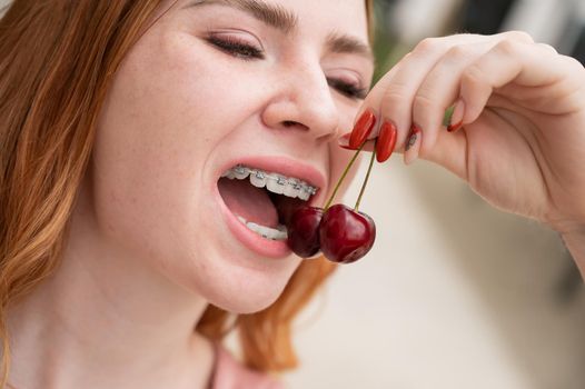 Beautiful young red-haired woman with braces on her teeth eats sweet cherries in the summer outdoors.