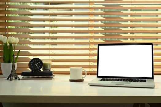 Laptop computer with white screen display, coffee cup, books and flower pot on white table.