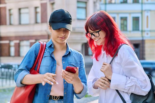 Two teenage females talking, looking into smartphone, outdoor on city street. Attractive girls friends, communicate together. Friendship, communication, adolescence, urban lifestyle, youth concept