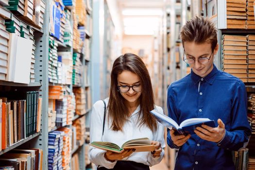 Two friends students reading books standing in the long corridor in library