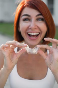 Red-haired Caucasian woman holding transparent mouthguards for bite correction outdoors. A girl with a beautiful snow-white smile uses silicone braces.