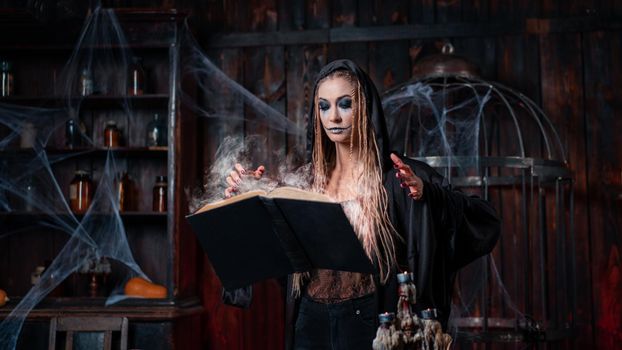 Halloween concept. Witch dressed black hood standing dark dungeon room use levitating magic book conjuring magic spell. Female necromancer wizard. Smoking spell book