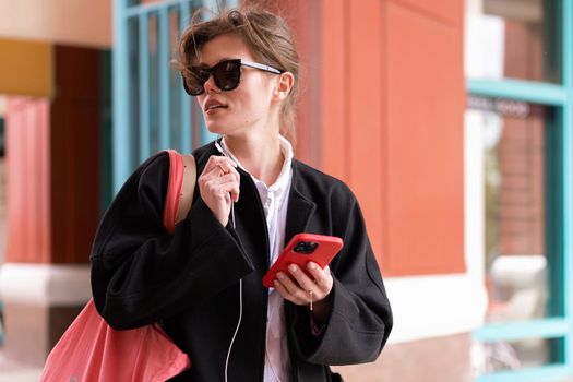 Portrait serious nice woman wearing black coat listening music with headphones and using smartphone. Beautiful caucasian female have phone conversation with earphones and mobile phone outdoor. Autumn