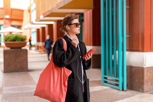 Portrait serious nice woman wearing black coat listening music with headphones and using smartphone. Beautiful caucasian female have phone conversation with earphones and mobile phone outdoor. Autumn