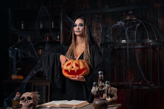Halloween concept. Black witch holding Halloween pumpkin with carved smily face in hand standing dark room. Female necromancer, dungeon place. Jack o lantern head. Wizard making scary curse
