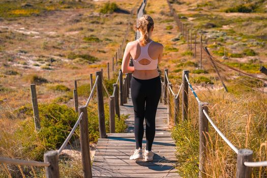 Athletic woman portrait outdoor. Caucasian female sportive woman walking wooden path way on nature ocean beach. Confident middle age fit woman summer day back view on sea beach Portugal