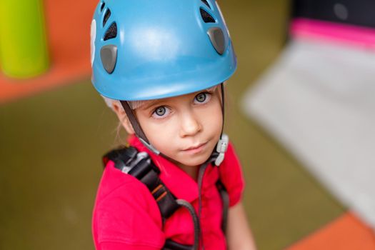 Cute little girl climber in blue protective helmet and gear for climbing standing in climber centre amusement park for children. Caucasian female climber 5 years
