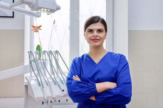 Portrait of confident female dentist in office looking at camera. Young female doctor in blue uniform with crossed arms, copy space. Dentistry, medicine, health care, profession, stomatology concept