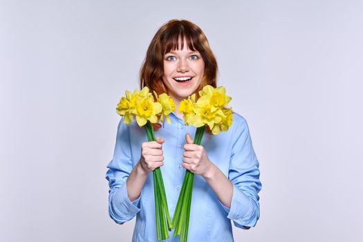 Portrait of young woman with bouquet of yellow flowers looking at camera, light background. Beautiful happy red-haired teenage female in blue shirt. Happiness, joy, holiday, beauty, youth concept
