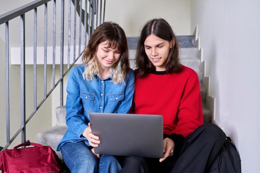 Teenage couple of friends students looking at laptop together. Teenagers male and female with backpacks sitting on the stairs inside the building. Youth, lifestyle, technology, young people concept