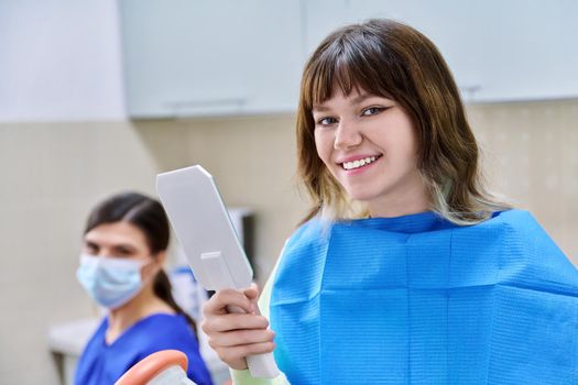 Teenage female looking at healthy teeth in mirror, in dental office, visit to dentist orthodontist. Treatment and care of teeth, teenagers, orthodontics, dentistry concept