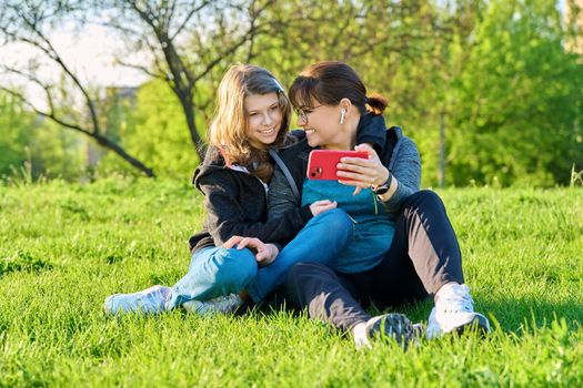 Mom and daughter 11, 12 years old having fun looking together at smartphone screen, outdoor, sitting embracing on grass. Family, leisure, lifestyle, relationship love, mother's day, motherhood concept