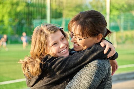 Portrait of happy mom and preteen daughter hugging together outdoor, near sports stadium, football field. Family, happiness, leisure, lifestyle, relationship, love, mother's day, motherhood concept