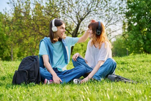 Couple of friends guy and girl 17, 18 years old sitting on grass. Students in headphones with backpacks sitting on campus lawn, talking and laughing. Teens, youth, friendship, education, young people