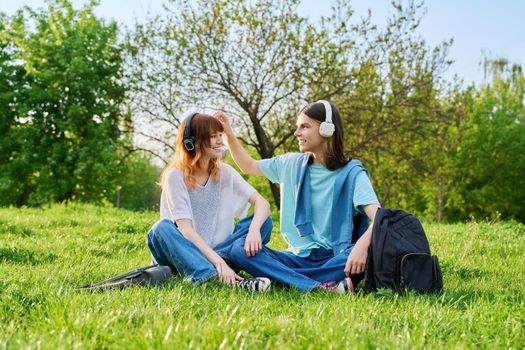 Couple of friends guy and girl 17, 18 years old sitting on grass. Students in headphones with backpacks sitting on campus lawn, talking and laughing. Teens, youth, friendship, education, young people