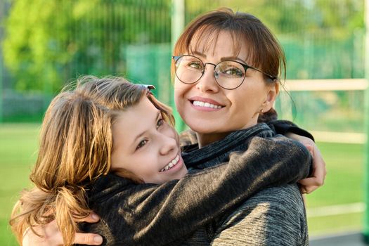 Portrait of happy mom and preteen daughter hugging together outdoor, near sports stadium, football field. Family, happiness, leisure, lifestyle, relationship, love, mother's day, motherhood concept