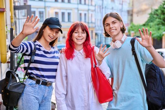 Group of teenage friends looking at the camera waving hand, outdoor. Three happy smiling teenagers on city street. Friendship, happiness, adolescence, urban lifestyle, education, students concept