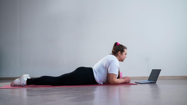 Young Plus Size Woman Stretching At Home Online. Flexible girl practices yoga and watches an online course on a laptop.